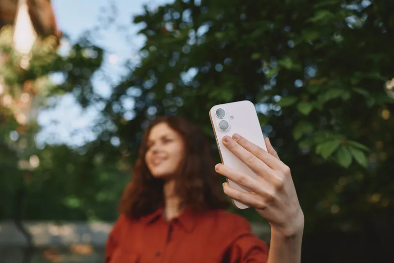 Candid woman lifestyle portrait taking a selfie outdoors, smiling softly in a red shirt, leafy park background, sunny day, relaxed casual mood, natural light