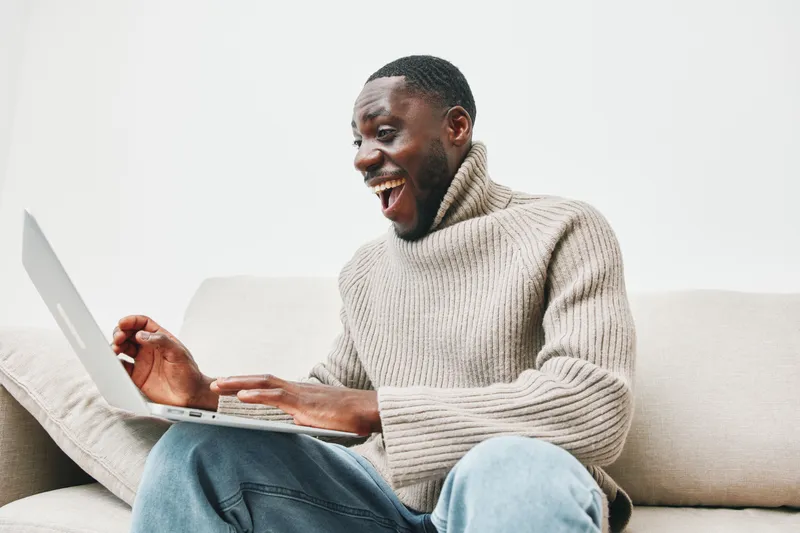 Happy Black man with a laptop enjoying online content in a cozy living room, wearing a beige sweater, evoking feelings of joy and comfort in a modern home setting