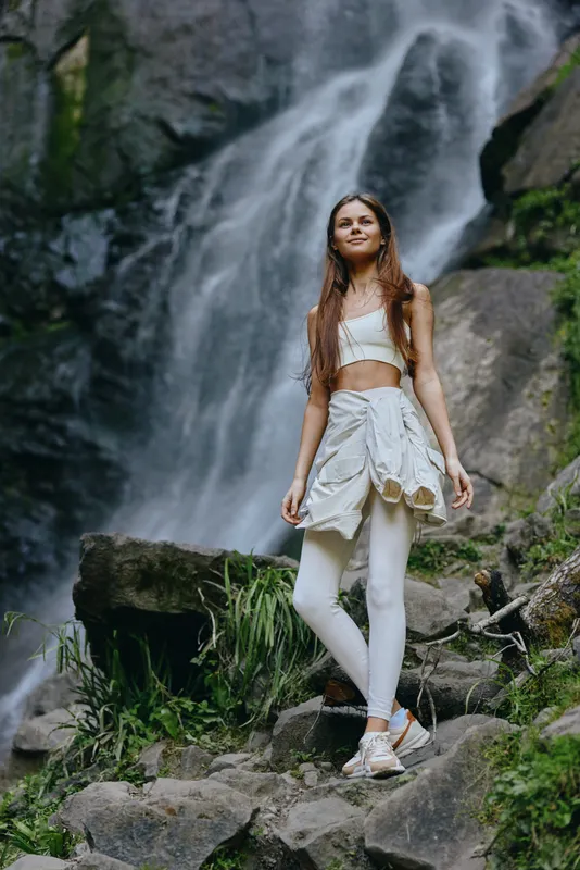 Young woman hiking near waterfall wearing white activewear and jacket tied around waist, surrounded by rocks and lush greenery in natural outdoor setting.