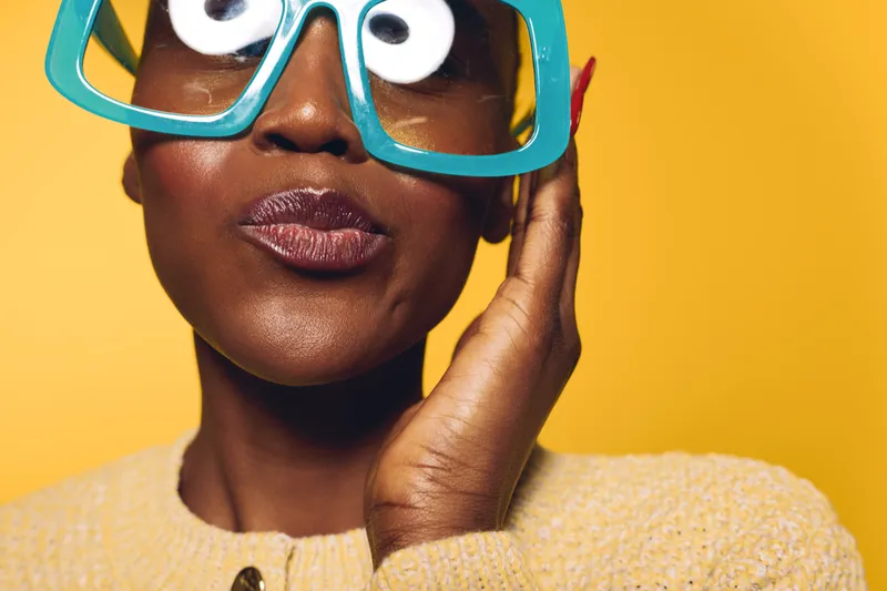 Close-up of dark skinned African woman wearing oversized blue glasses and yellow sweater against yellow background. Concept of fashion, style, beauty, and playful expression.