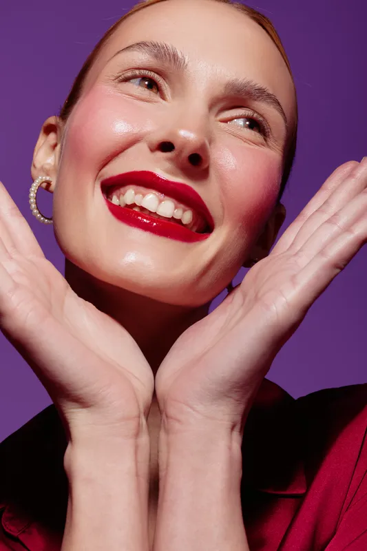 Close-up of happy woman with red lipstick smiling and holding hands near face on purple background. Concept of beauty, makeup, joy, and positive emotions in studio lighting.