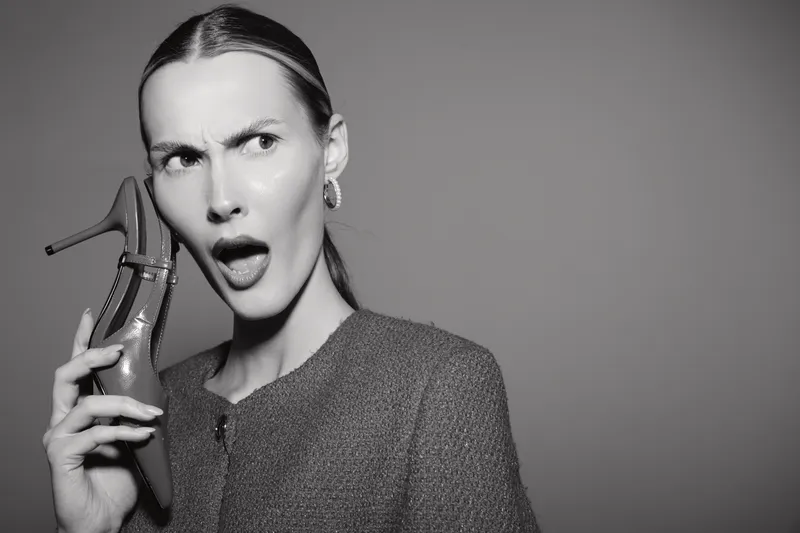 A young white woman with pale skin and slicked back hair holds a high heel shoe near her ear, expressing surprise or confusion in a monochrome studio setting.