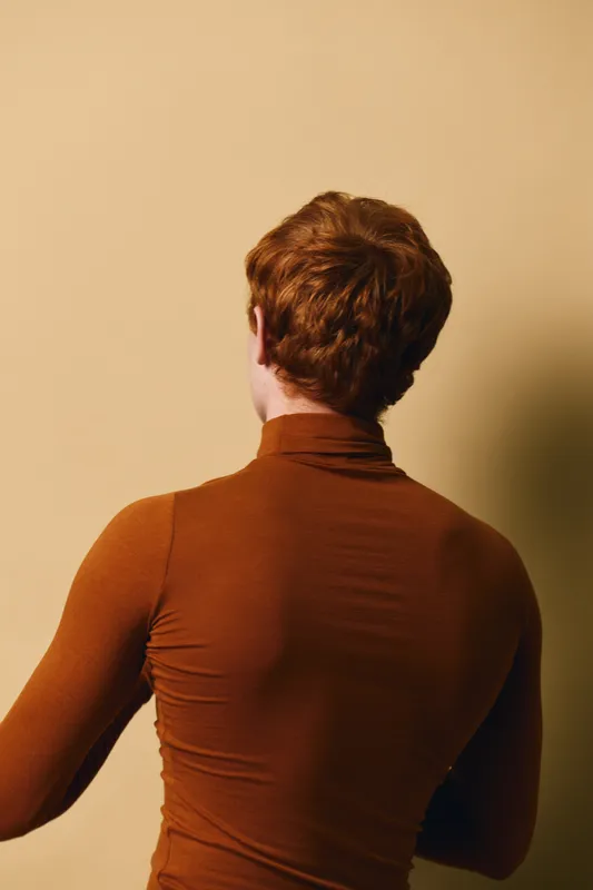 Person with red hair wearing brown turtleneck standing with back to camera on beige background in studio for fashion portrait concept and casual style.