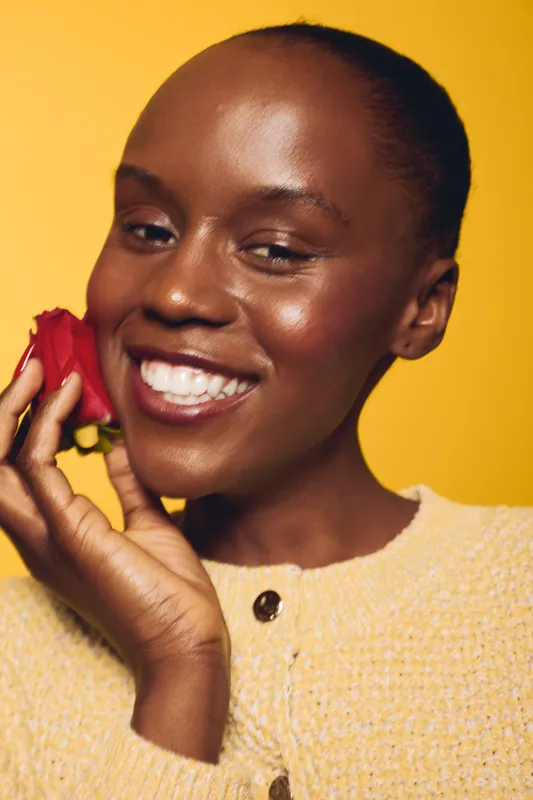Close-up of happy black African woman with dark skin holding red rose near face smiling on yellow background. Concept of beauty, skincare, natural glow, and positive emotions.