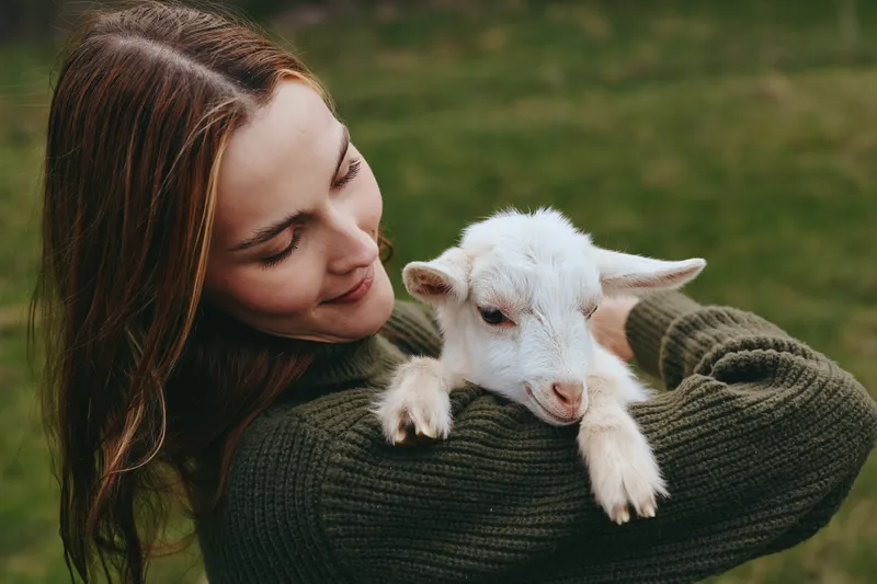 A woman in a dark sweater holds a goat kid in a lush countryside pasture, sharing affection and gentle contact. An outdoors scene that conveys animal bond, comfort, and wholesome lifestyle.