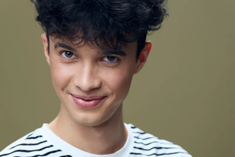 young man smiling close-up portrait wearing striped shirt with curly hair and warm beige background revealing friendly and confident expression