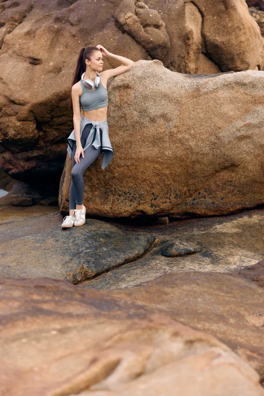 Young woman in activewear on rocks by the beach, showcasing fitness and vitality with a serene expression and natural backdrop