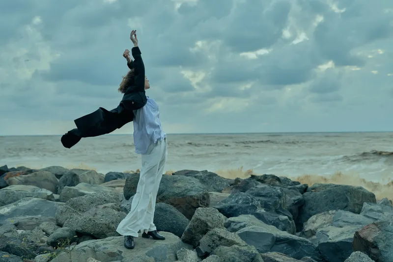 Dancer on rocky shore performing with arms raised, waves crash nearby, wind swept motion against a cloudy sky and expansive sea backdrop