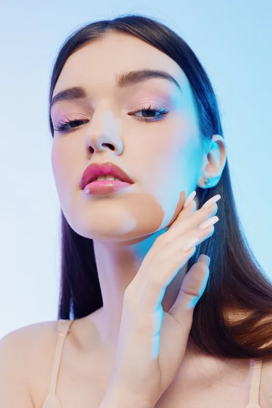 Young woman beauty fashion model with long hair poses with hand near face. Soft studio lighting highlights her flawless skin and glossy lips. Concept of elegance and skincare.