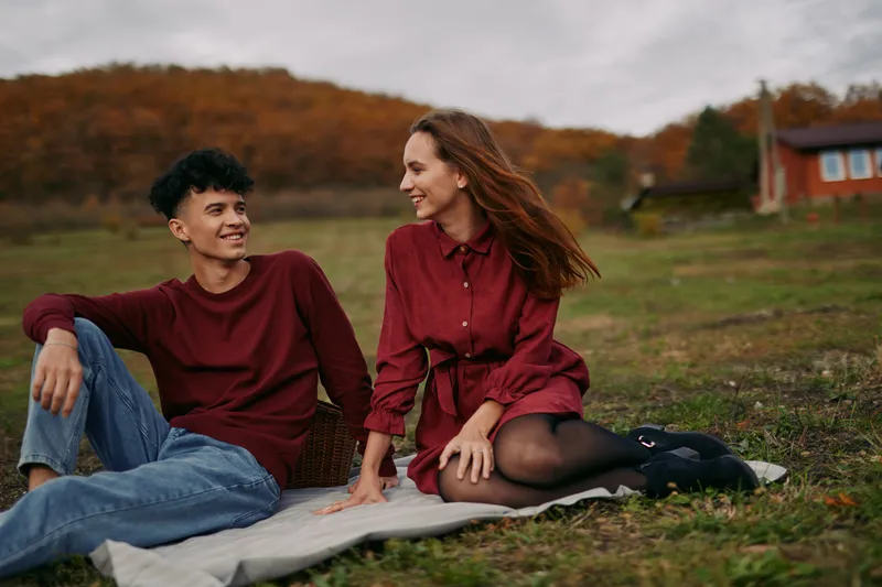 A candid outdoor portrait of a young couple sitting on a blanket in a meadow, sharing a genuine smile and relaxed body language that conveys authenticity and calm connection.