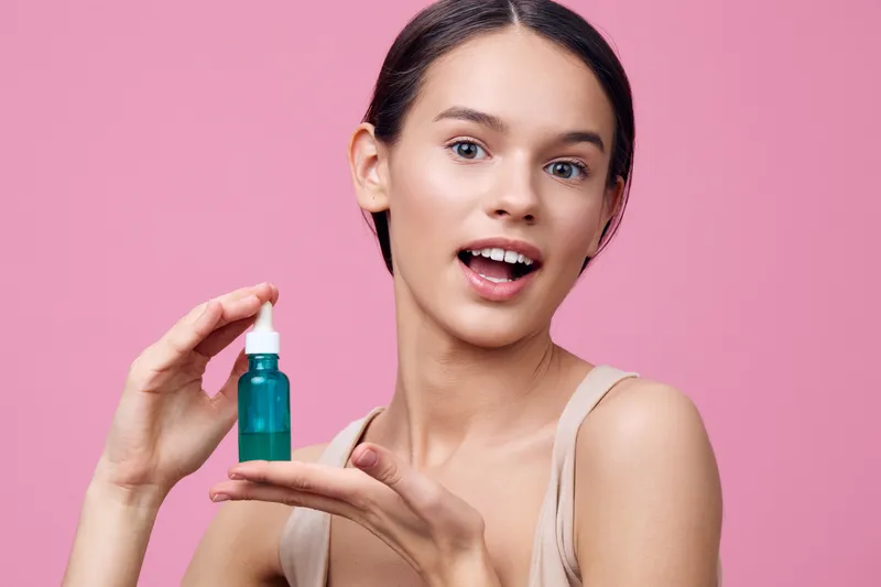Happy young woman holding a green skincare serum bottle against a pink background, showcasing healthy skin and vibrant beauty, ideal for cosmetics marketing