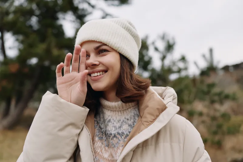 woman outdoors smiling and waving hand in a cozy beanie and light coat in a natural setting, capturing candid winter fashion and casual mood with soft background scenery