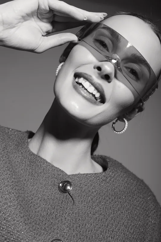 Smiling caucasian woman with light skin wears large sunglasses and hoop earrings, dressed in textured sweater. Studio portrait with stylish and happy mood.