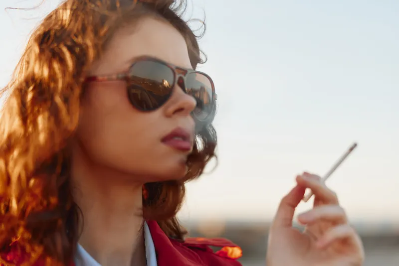 Young woman with curly hair wearing sunglasses smokes cigarette outdoors during sunset with cityscape background in soft warm light and casual red jacket.