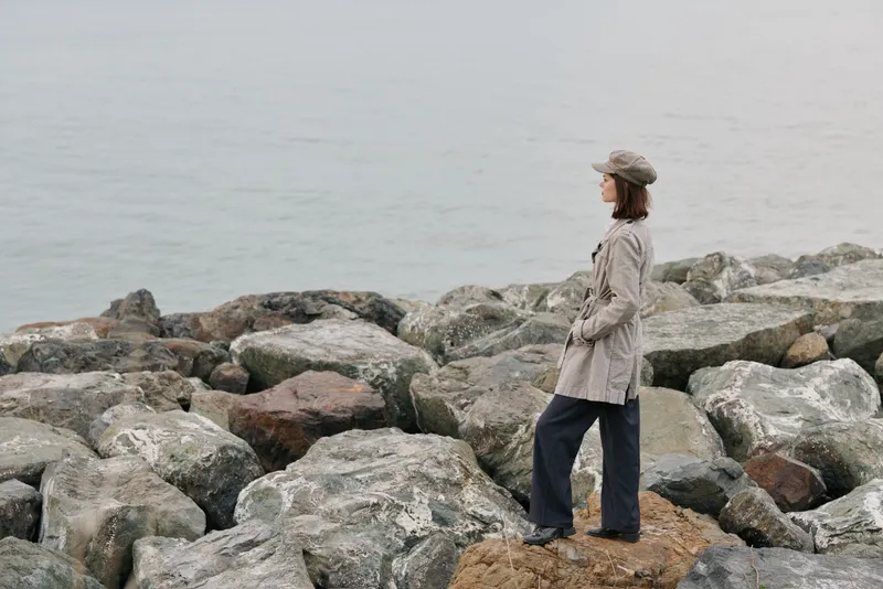 Woman in plaid coat and hat stands on rocky shore looking at calm sea. Overcast day with muted colors creates peaceful mood of contemplation and nature connection by water.