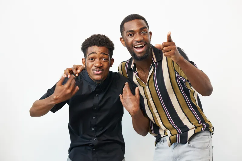 Two black men sharing a joyful moment, displaying smiles and playful gestures in a bright setting. Their vibrant outfits enhance the lively atmosphere captured in this image.