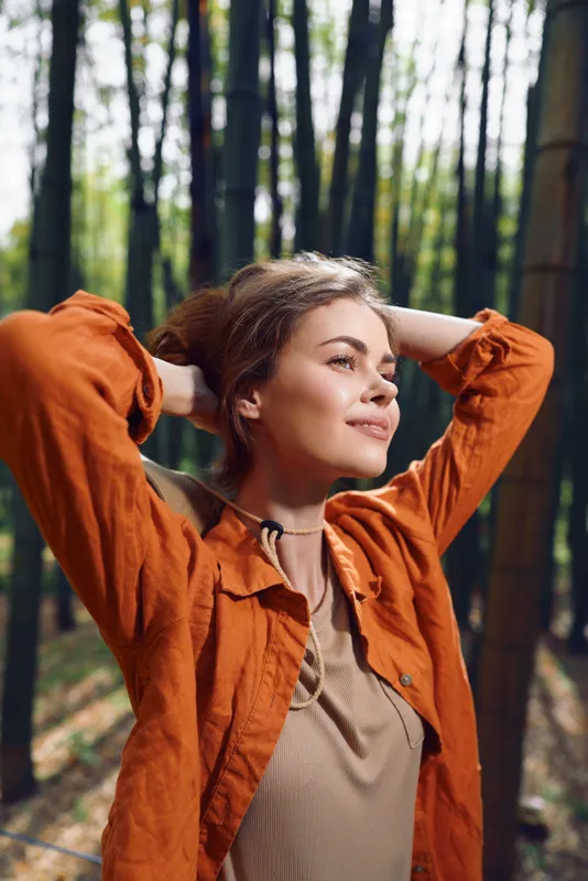 Woman portrait in a forest enjoying nature and sunlight, relaxed outdoors among tall trees. Casual jacket and content smile, peaceful mood for wellness, mindfulness and leisurely escape.