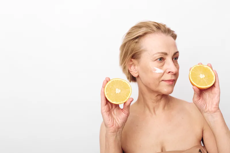 Mature woman with short hair holds two halves of fresh lemon near face with white cream on cheek. Concept for natural skincare, vitamin c, and healthy aging beauty routine.