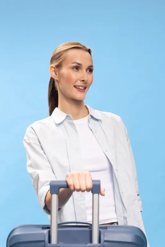 Young woman with long hair smiles while holding suitcase handle, wearing casual white shirt on blue background. Concept for travel, vacation, tourism, and leisure.