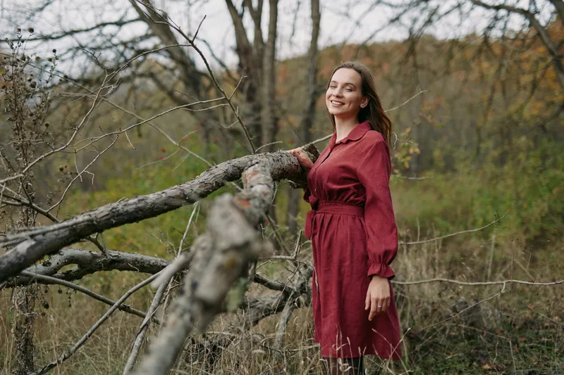 A candid portrait of a woman in a burgundy dress near a fallen log in a rural field, radiating authenticity and credibility through a natural, unposed moment.