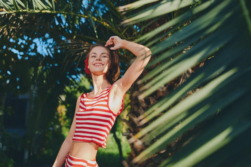 A cheerful woman wearing a red and white striped swimsuit stands outdoors among lush greenery, smiling brightly in a sunny tropical scene.