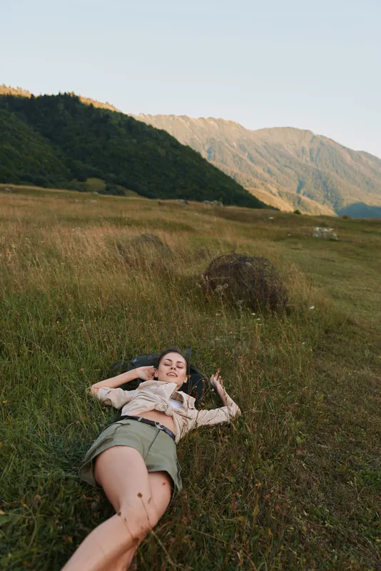Relaxed woman lying in a grassy field with distant mountains, capturing a carefree outdoor lifestyle moment. Soft natural light creates a warm, tranquil mood and well being.