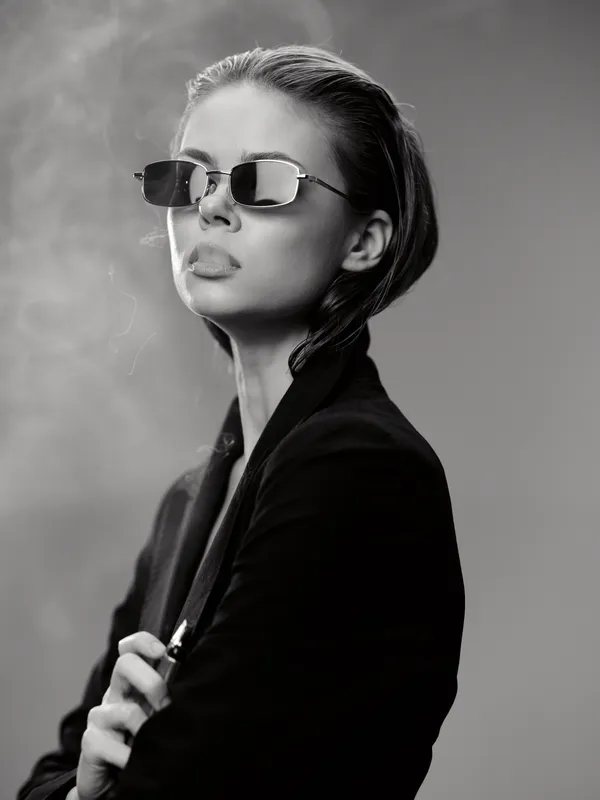 Stylish woman wearing sunglasses and smoking, black and white portrait with confident expression.
