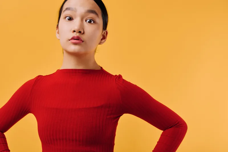 Young woman in red sweater standing with hands on hips against orange background looking surprised or curious with expressive face and confident pose in studio.