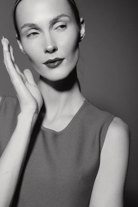 Portrait of a slender white woman with pale skin and slicked hair in a sleeveless top, posing with hand near face against dark background. Concept of elegance and minimalism.