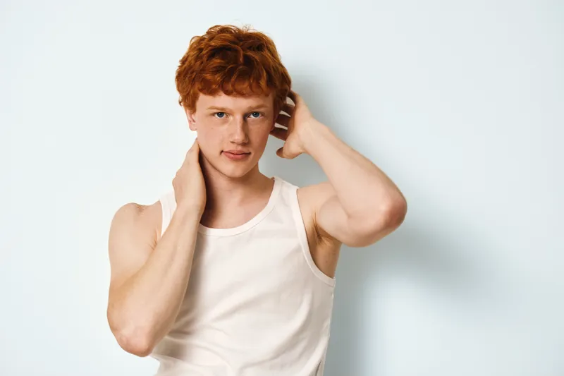 Young man with red hair and fair skin posing against light background wearing white sleeveless shirt with hand touching neck in casual style portrait expression.