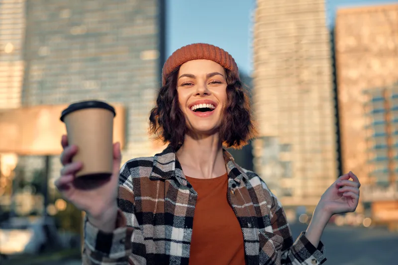 Woman holding coffee cup and smile in urban street at golden hour glow, candid beanie portrait showing authenticity, mindful living and emotional storytelling in a modern city lifestyle.