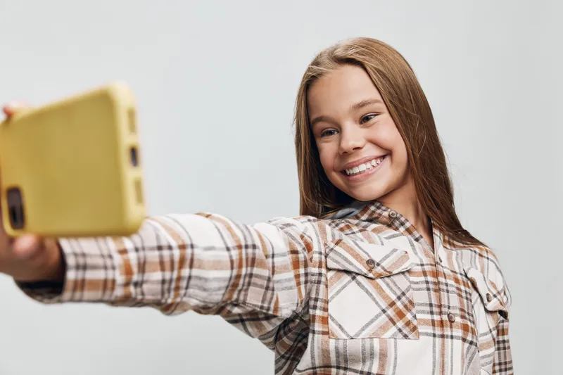 Teenage girl with long hair in plaid shirt taking selfie with yellow smartphone smiling and posing on neutral background for social media or personal photo session.