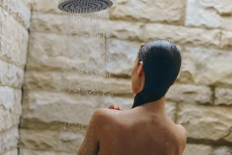 Woman showering outdoors under rain showerhead with natural stone wall background, wet hair, and water droplets flowing down her bare back, peaceful moment of self-care.