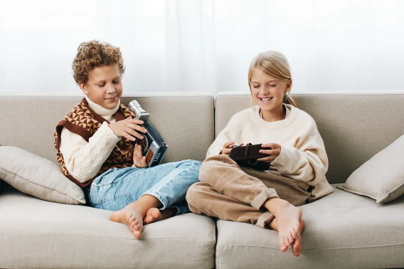 Authentic lifestyle scene of school age siblings enjoying gaming on a cozy sofa; one holds a handheld console, the other a controller, captured in a natural, candid moment.