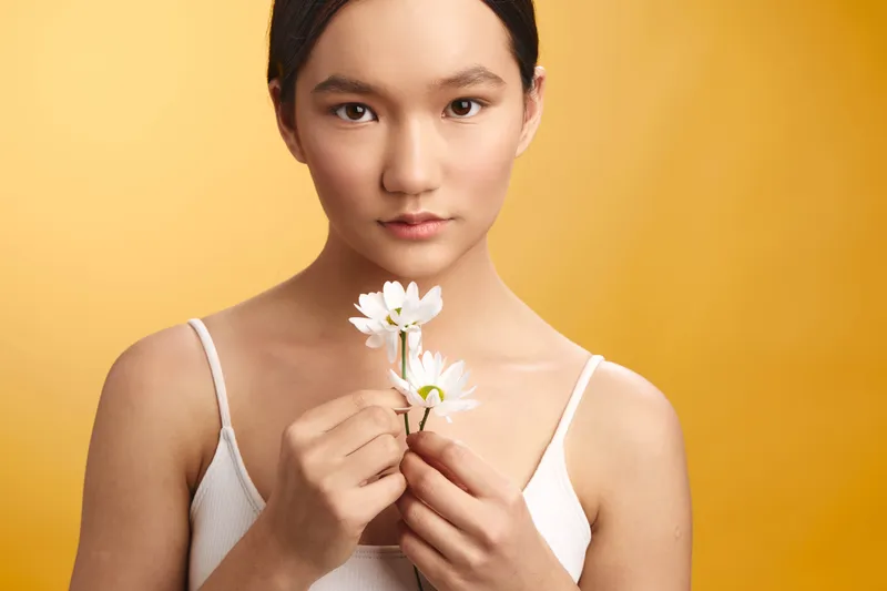 asian woman wearing a white tank top holds delicate flowers against a warm orange backdrop, gazing softly at the camera to convey calm elegance and a serene studio portrait.