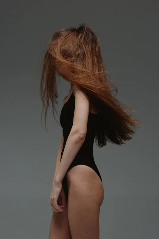 woman with long red hair posing in black bodysuit with flowing hair and minimalistic grey background