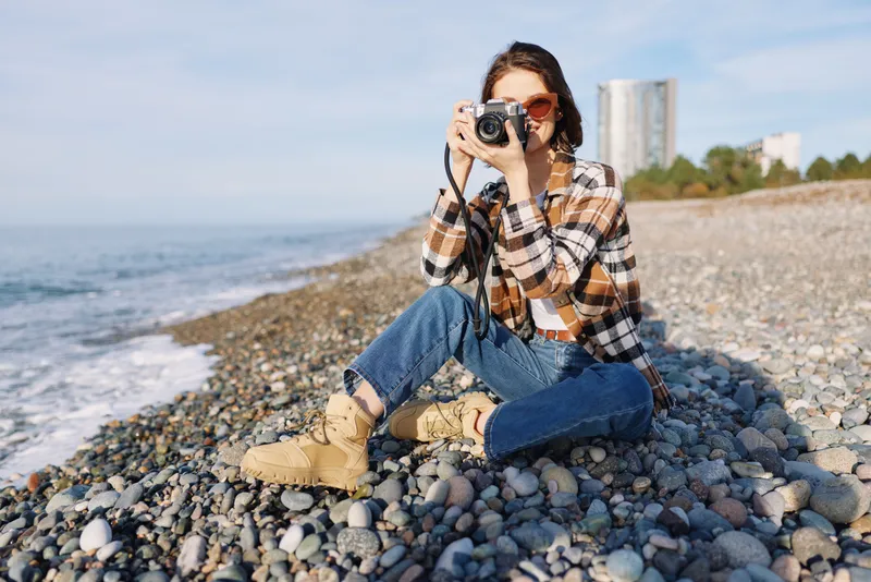 Woman photographer on a pebble beach with a camera, wearing a plaid shirt, jeans and boots, capturing coastal scenery and bright outdoor moments by the sea