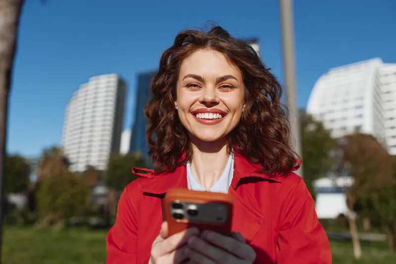 Young woman with curly hair smiles joyfully while using a smartphone outdoors in a city park on a sunny day.