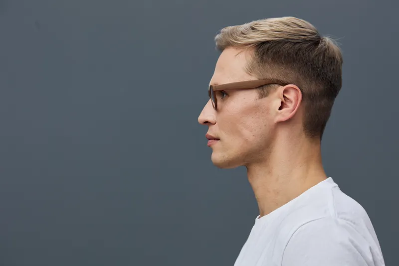 Portrait of a young man in profile view with glasses, wearing a white t-shirt, against a plain gray background. Studio shot capturing calm expression, modern style, minimalistic concept, suitable for 