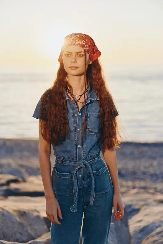 Lifestyle woman in modern boho-western denim street style with a red bandana, standing outdoors near the sea at sunset in warm film color tones.