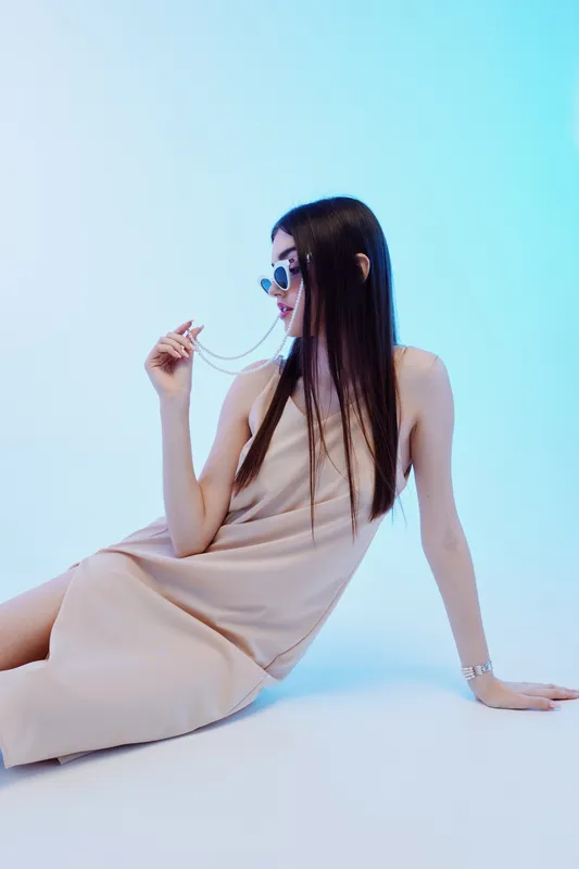 Young woman in a beige dress poses seated on a white floor against a light blue background. She wears sunglasses and holds a necklace, showcasing fashion and beauty concepts.