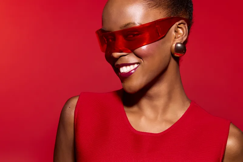 Happy dark skinned African woman in red dress and futuristic red glasses smiles against red background. Concept of fashion, beauty, style, and confident female model portrait.