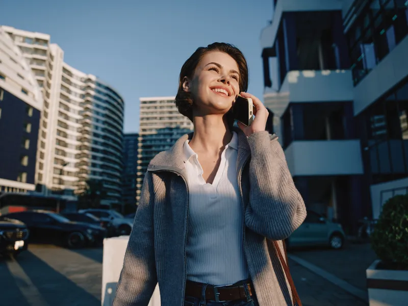 A cheerful professional woman is seen talking on a phone in a modern urban environment. She smiles, exuding confidence and positive energy in a bright outdoor setting.