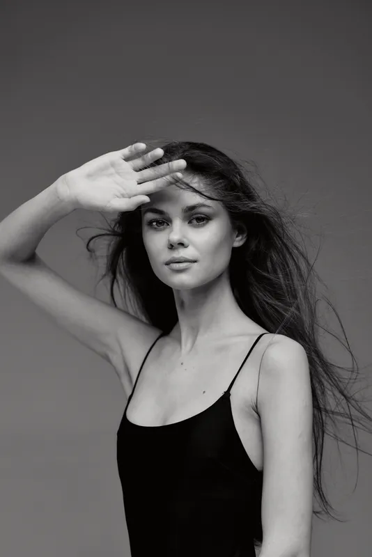 Black and white portrait of a young woman shielding her eyes with hand, wearing black sleeveless top with loose hair