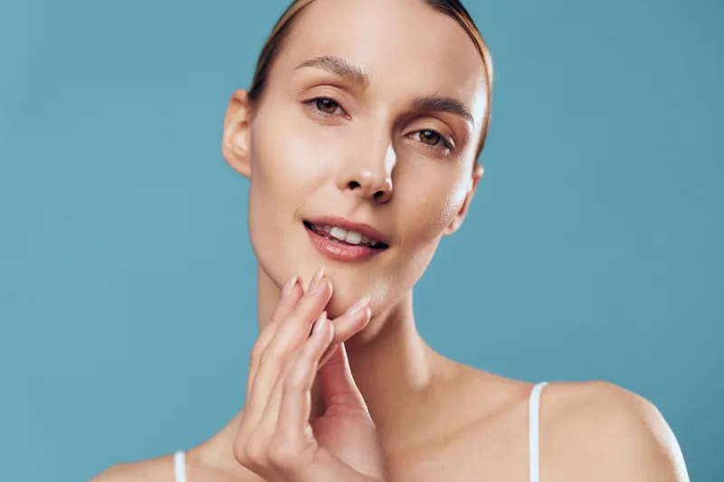 Young woman with clear skin gently touching her face against blue background. Concept of skincare, beauty, wellness, and natural care. Soft lighting highlights healthy complexion and calm expression.