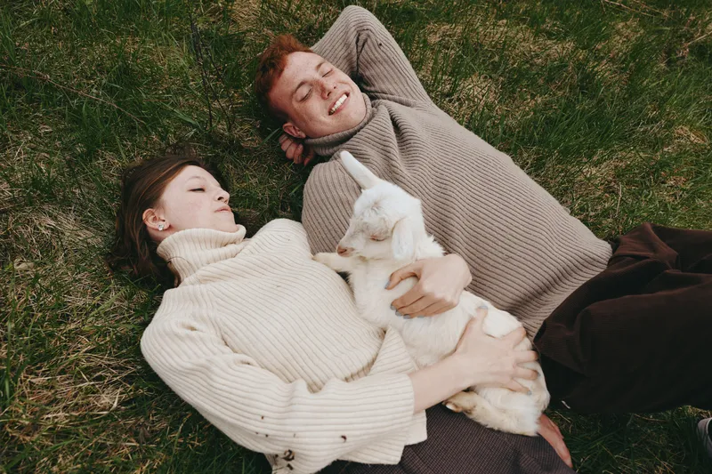 Couple lies on grass enjoying a cozy moment outdoors with a friendly goat kid between them, conveying simple lifestyle intimacy amid a natural rural setting and calm countryside mood.