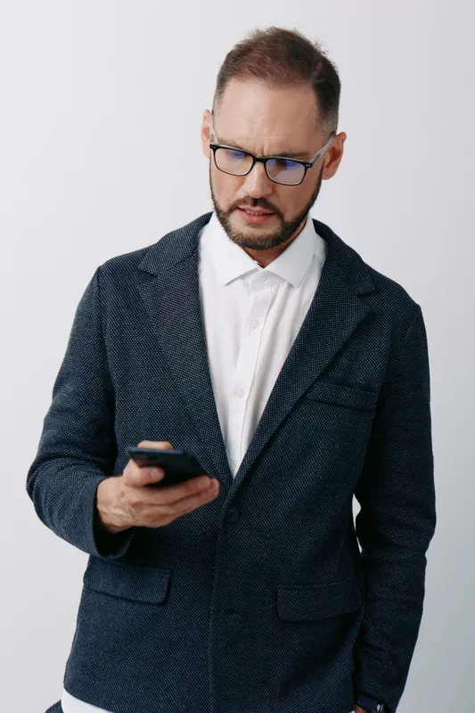 A focused business professional stands against a solid colored backdrop, examining a phone with a thoughtful, attentive expression. The scene conveys quiet confidence, preparedness, and calm control.