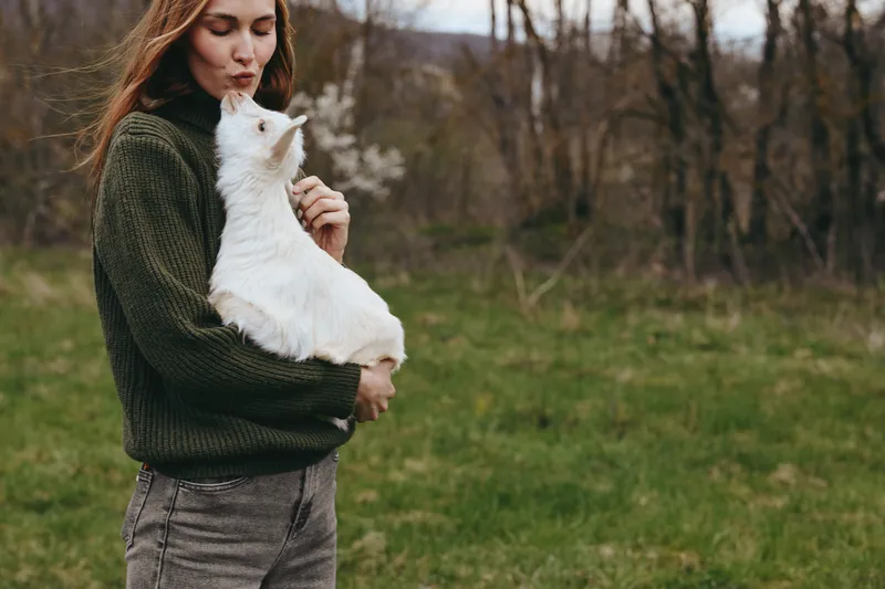 A woman in a knitted sweater cradles a white goat kid outdoors in a grassy field, sunlight filtering through trees. A gentle moment of bonding, rural life, and animal companionship in natural light.