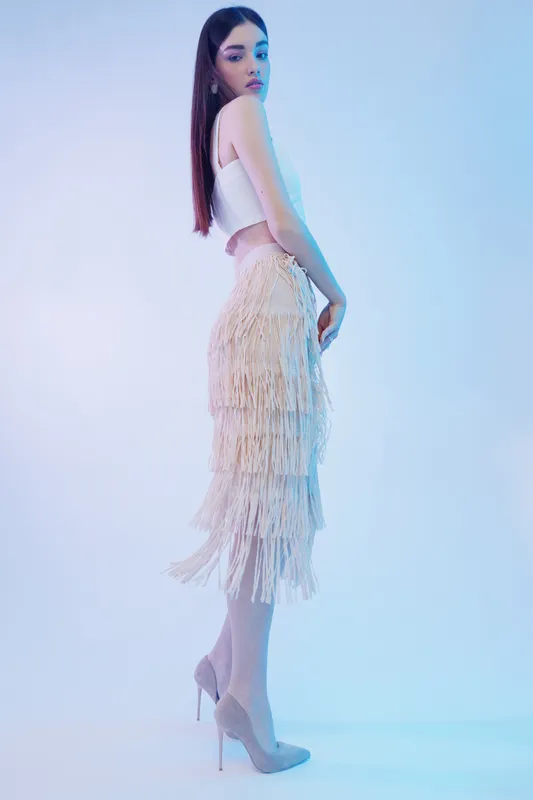 Young woman posing in a stylish fringe skirt and white crop top. She wears high heels and looks confidently to the side. Studio shot with soft blue lighting. Concept of woman beauty fashion.