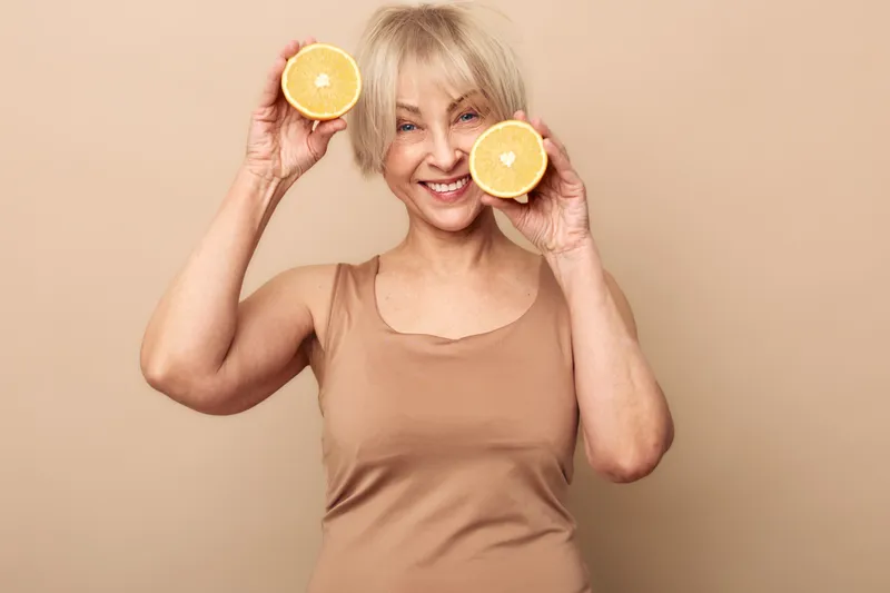 Smiling mature woman with short blonde hair holds two halves of fresh orange near face on beige background. Concept of healthy lifestyle, vitamin C, natural skincare and wellness.